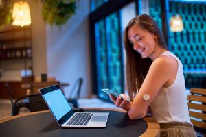 A smiling woman with a round sensor on her arm checks her smart phone while sitting at a cafe table with a laptop open in front of her.